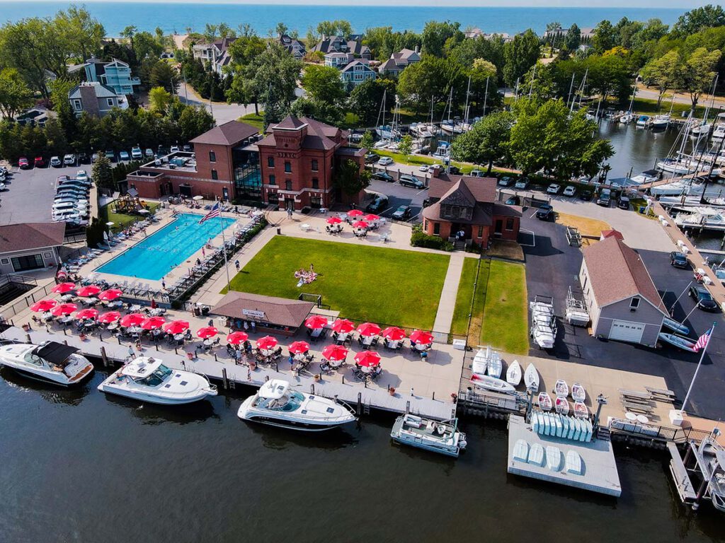 Aerial view of the St. Joseph River Yacht Club in St. Joseph, Michigan showing a large pool, a large green space, two large brick buildings, several red umbrellas  and boats along the shore. 