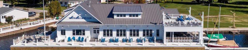 Aerial view of the Pentwater Yacht Club in Pentwater, Michigan. White building with a gray roof on a dock with many blue deck chairs in front