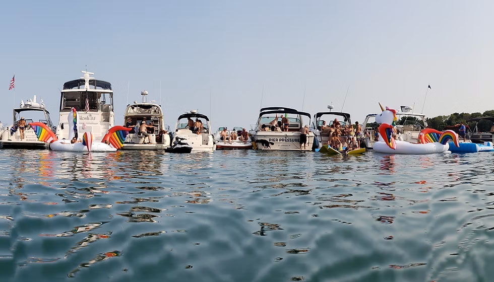 A line of boats are shown side by side in a festive atmosphere, with lots of large floaties and people all around having fun on the water