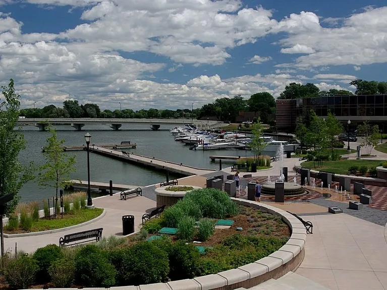 A beautiful harbor is seen against a blue sky with fluffy clouds. There are boats docked along the shore, and a beautiful waterside walkway with landscaping and a small water play area with ground fountains.