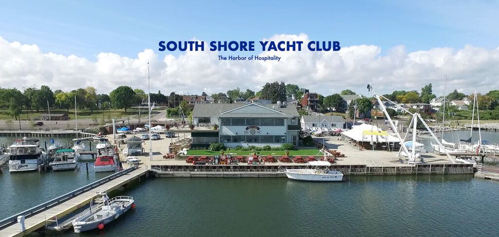 Aerial view of the South Shore Yacht Club from the water. Boats surround the dock, where the SSYC building is seen in the center, a two-story white building with "SSYC" on the front.