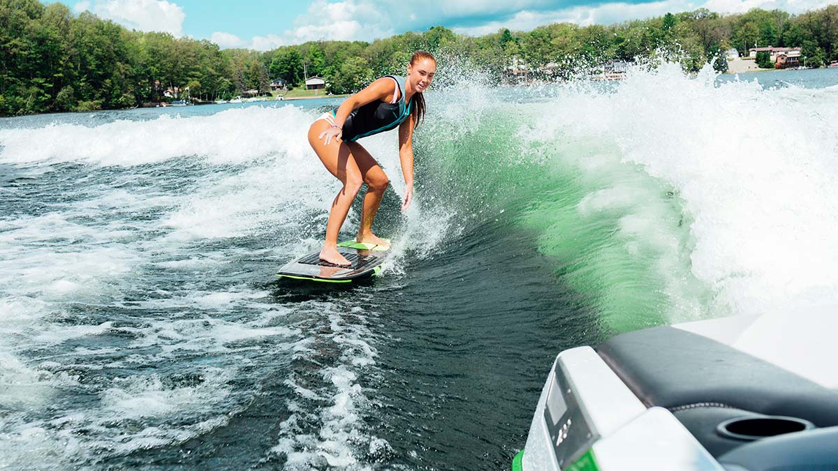A woman in a black and teal wetsuit wakesurfs on a large, cresting green wave behind a boat. She rides a short board without a tow rope. The view is from the boat's stern, looking back at the churning white wake and a tree-lined shoreline with houses.